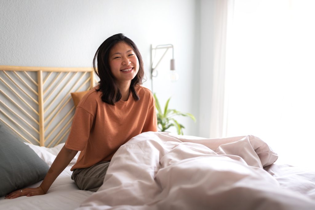 Happy girl sitting on bed waking up in the morning in cozy bedroom looking at camera. Copy space.